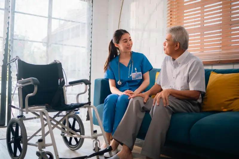 Nurse talking with an older man on a sofa, wheelchair beside them