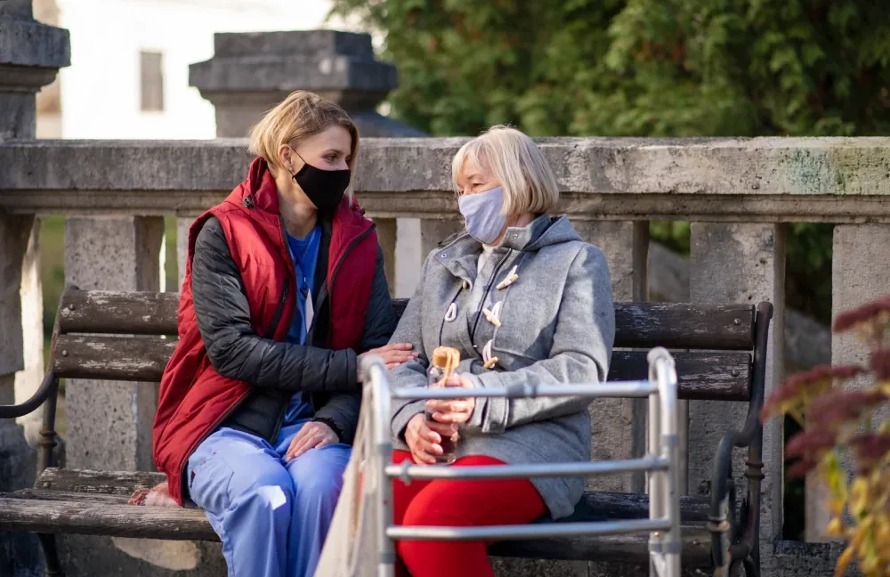 Support worker and older woman with a walking frame sitting outdoors, both wearing masks