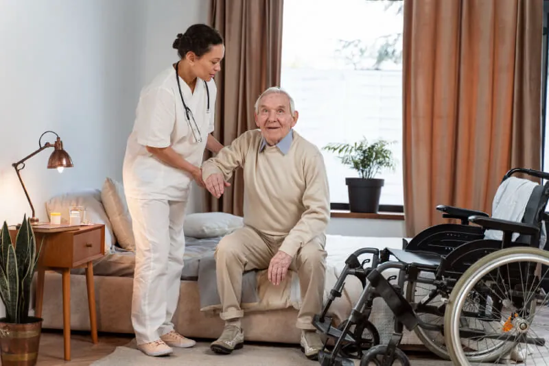 Support worker helping an older man to stand from a sofa, wheelchair beside them