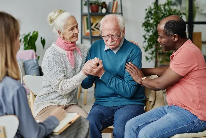 Older man holding hands with family members and a support worker in a living room