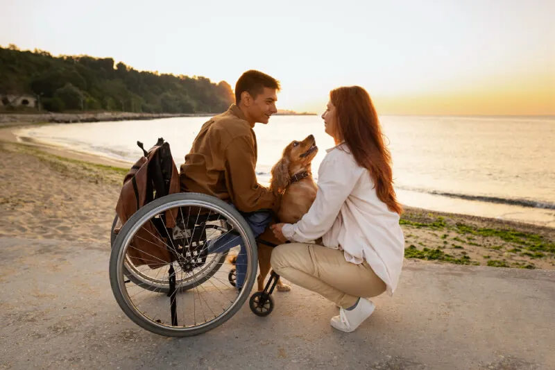 Man in a wheelchair with a companion and a dog on the beach at sunset