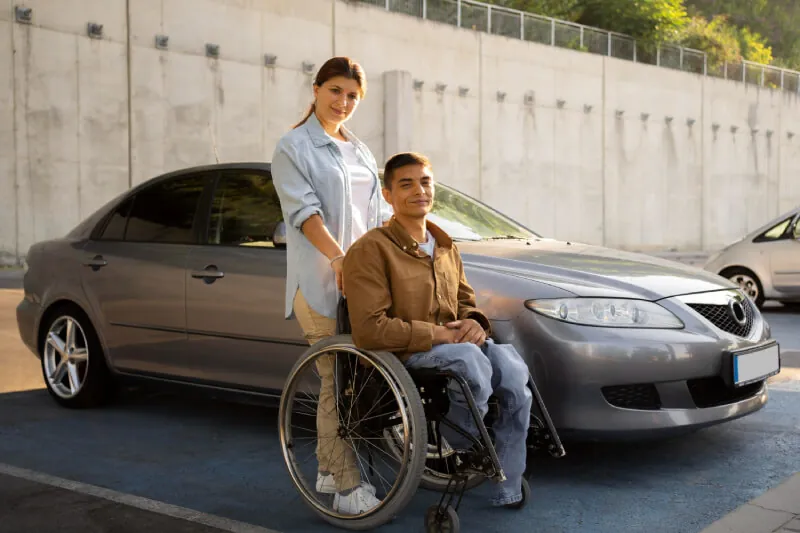 Support worker standing with a man in a wheelchair beside a parked car