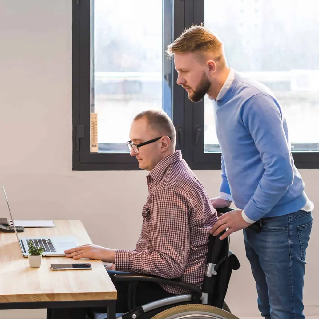 Man in a wheelchair working at a laptop with a colleague looking on