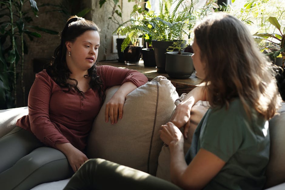 Two women having a heartfelt conversation on a cozy sofa surrounded by green plants indoors.